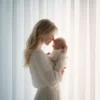 Mama w białej sukni tuląca noworodka przy oknie symbol bliskości, spokoju i opieki po porodzie.